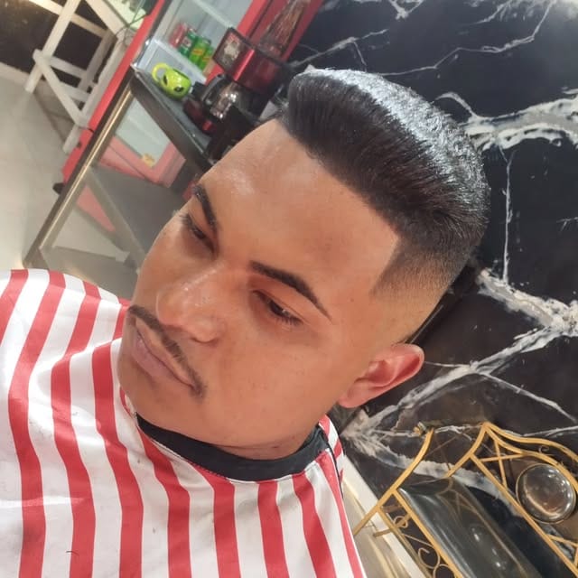 Cliente com topete e degradê nas laterais, sentado na cadeira em frente à parede de mármore preto